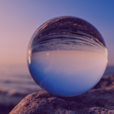 Picture of crystal ball perched on a rock with the ocean in the background. All the imagery is a muddled reflection within the ball.